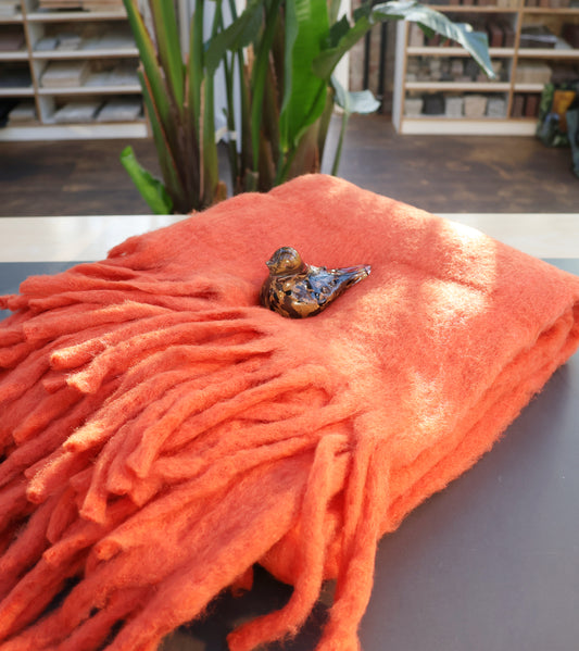 Die Decke aus Wollmischung | orange von Werner Voss liegt gefaltet auf einem Tisch, darauf steht ein dekorativer Keramikvogel. Im unscharfen Hintergrund sind grüne Pflanzen und Bücherregale zu sehen.