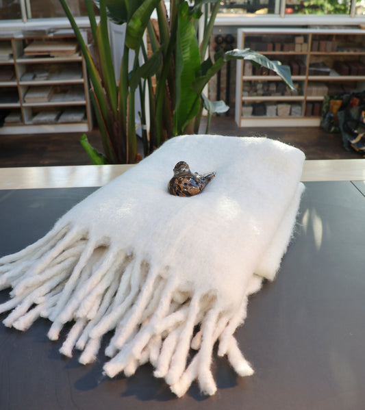 Die weiße Decke aus Wollmischung von Werner Voss ist auf einen Tisch gefaltet, ein kleiner Keramikvogel sitzt darauf; im Hintergrund sind Pflanzen sowie Regale mit Büchern und Materialien zu sehen - gemütliches Ambiente.