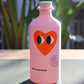 Eine rosafarbene 500-ml-Flasche Natives Olivenöl - Love Edition von yiayia and friends, auf deren Vorderseite ein rotes Herz mit zwei Augen abgebildet ist, steht auf einem Tisch mit unscharfen grünen Pflanzen im Hintergrund.