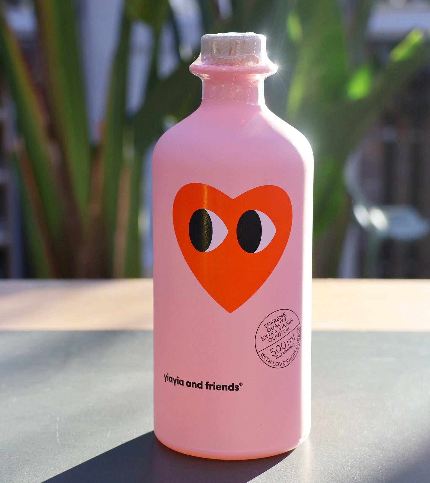 Eine rosafarbene 500-ml-Flasche Natives Olivenöl - Love Edition von yiayia and friends, auf deren Vorderseite ein rotes Herz mit zwei Augen abgebildet ist, steht auf einem Tisch mit unscharfen grünen Pflanzen im Hintergrund.