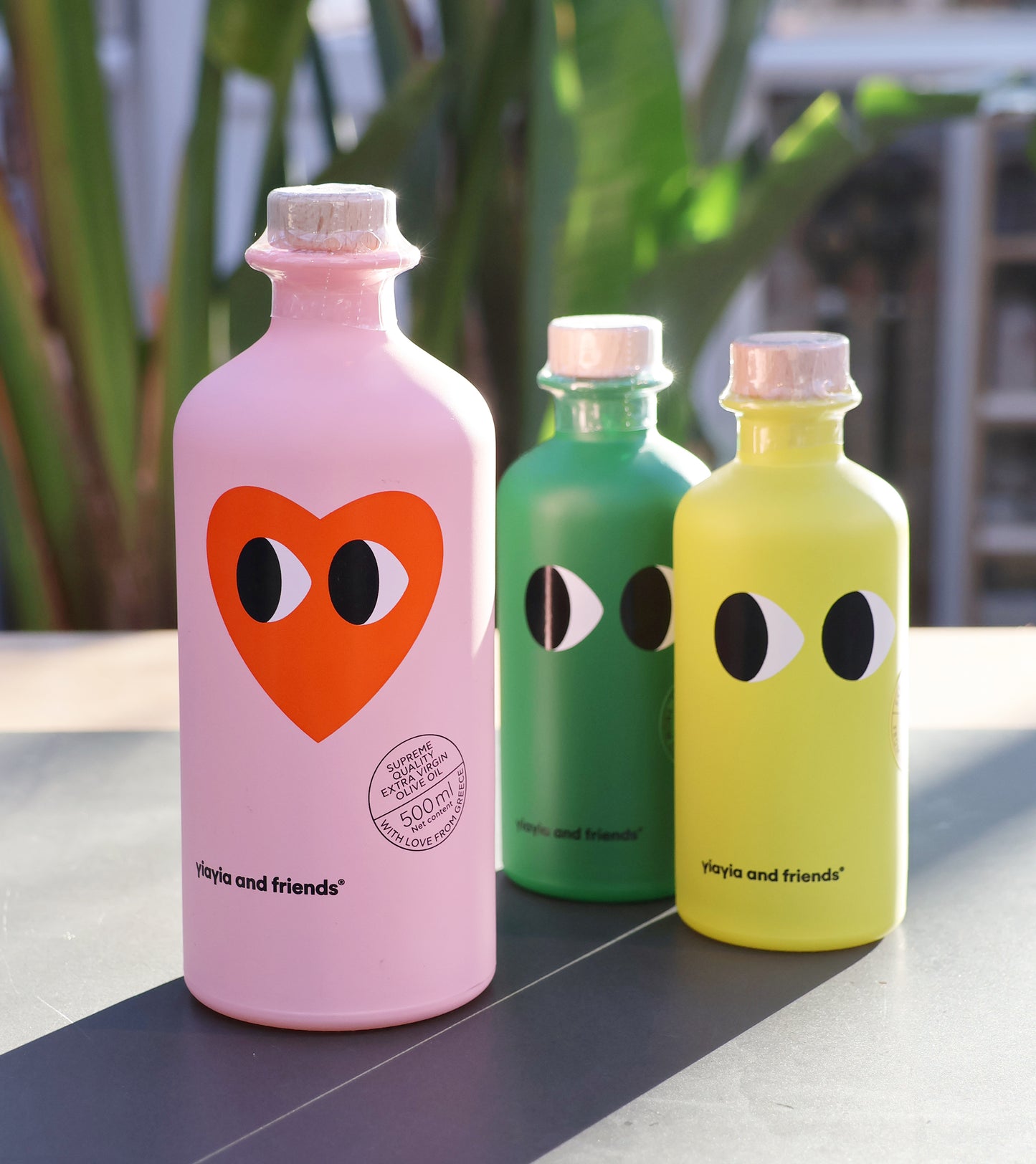 Drei farbenfrohe yiayia and friends-Flaschen sind mit verspielten Augenmotiven und dem Text "yiayia and friends" versehen. Die rosa Natives Olivenöl - Love Edition 500 ml Flasche hat ein großes orangefarbenes Herz mit Augen; grüne und gelbe Flaschen zeigen einfache schwarz-weiße Augen. Dahinter erscheinen Pflanzen.