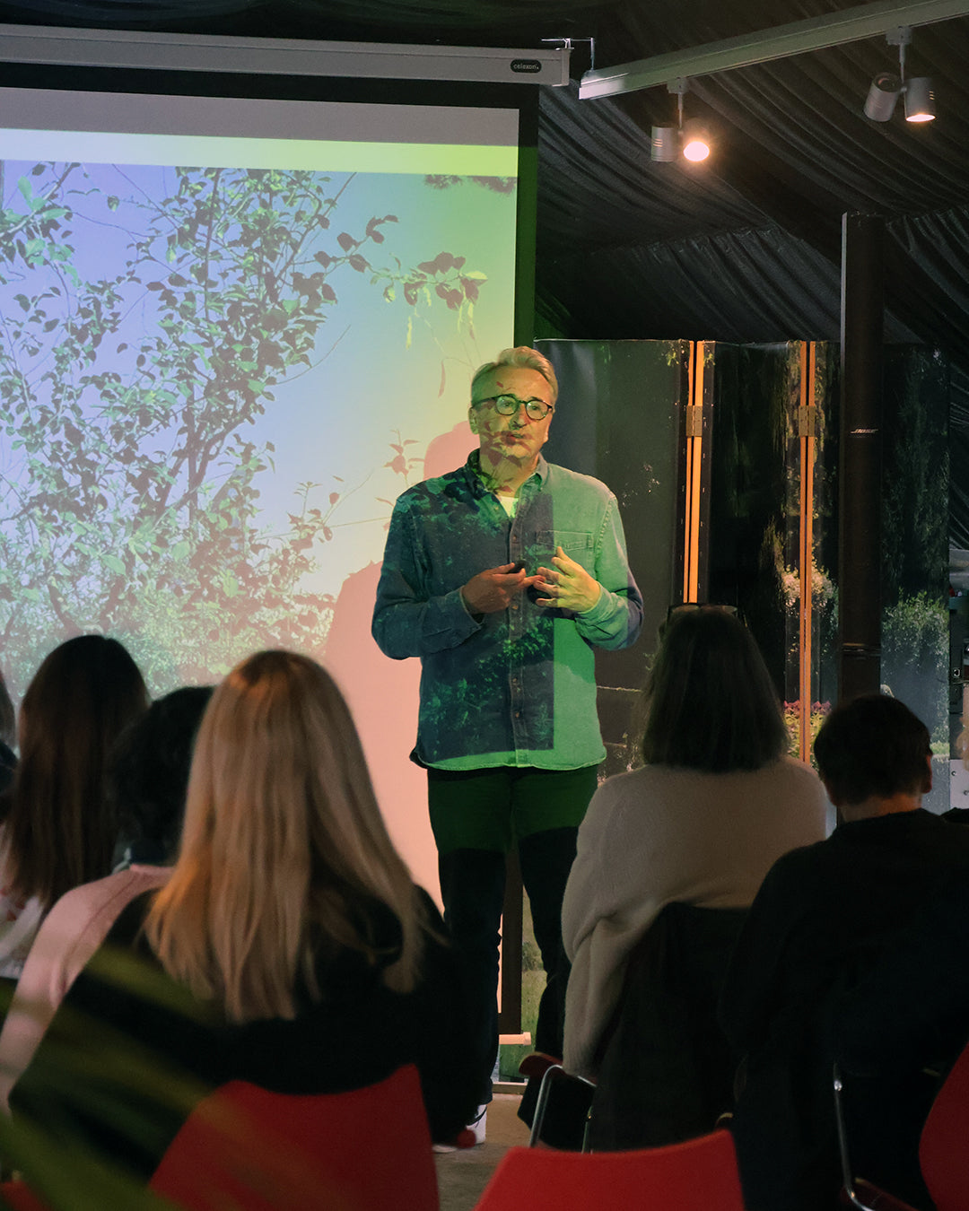 Eine Person präsentiert "Fr. 13.03.2026 | 13.00-16.00 HALBTAGESKURS Das Gartenjahr-Monat für Monat mit Horst Mager" von Horst Mager, mit einer Naturszene im Hintergrund; das Publikum sitzt auf roten Stühlen unter Scheinwerfern in einem dunkel getäfelten Raum.
