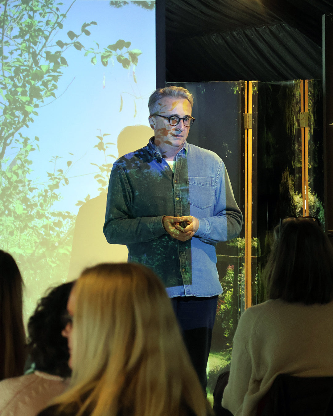 Horst Mager, mit Brille und Jeanshemd, fesselt in seinem Halbtageskurs "Das Gartenjahr-Monat für Monat" (Fr. 13.03.2026 | 13:00-16:00) ein aufmerksames Publikum, hinter dem er eine üppige Gartenjahr-Szene projiziert - ein inspirierender Moment.