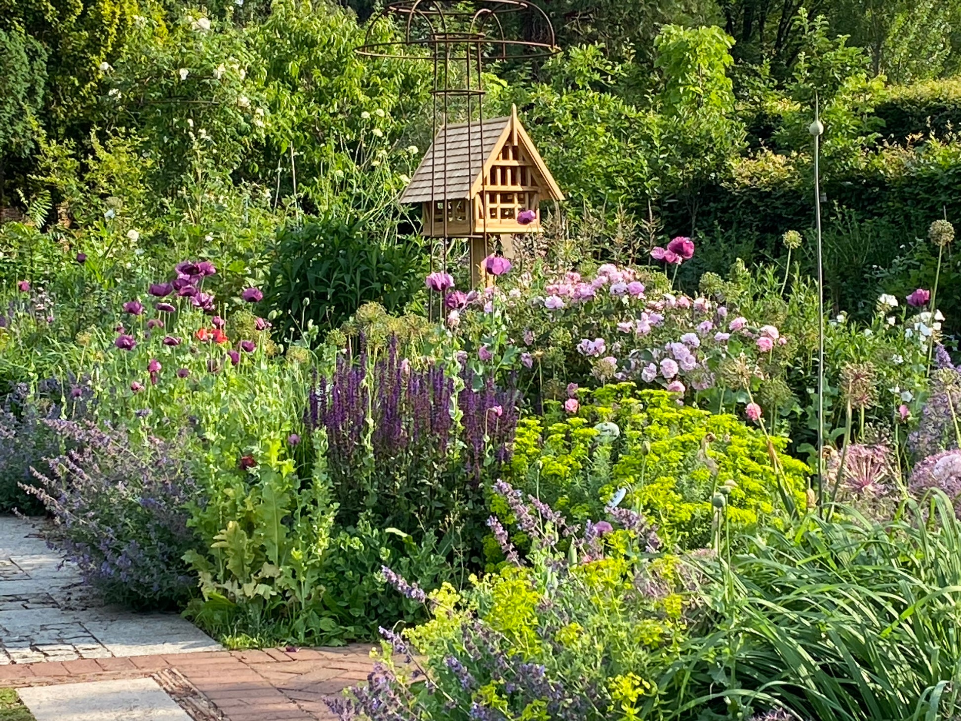 Ein üppiger Garten mit bunten Blumen und hohem Grün unterstreicht die fachkundige Gartenpflege. Ein hölzernes Vogelhäuschen steht neben einem Weg, der an lebendige Staudenbepflanzung grenzt - Fähigkeiten, die in der Meisterklasse Gartenpflege der Königlichen Gartenakademie (03.-04.02.2026) erlernt werden.