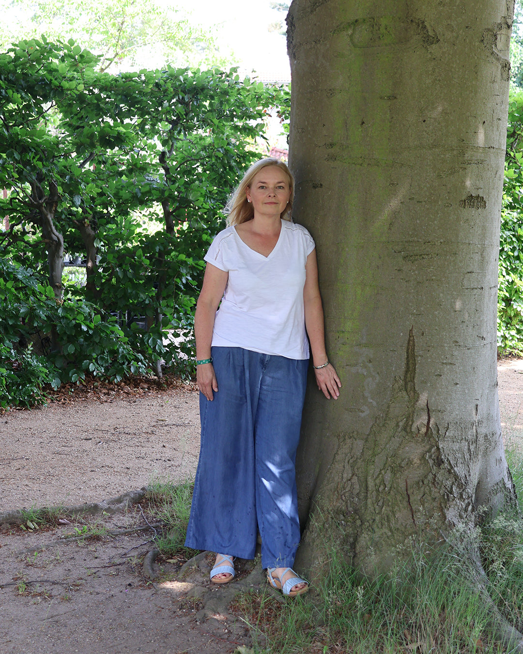 Eine Frau mit langen blonden Haaren, in weißem Hemd und blauer Hose, steht an einem Baum - eine einladende Szene für alle, die sich für die Fr. 27.02.2026 | 09.30-12.30 HALBTAGESKURS Grundkurs "Grüner Daumen" von Astrid Torrens.