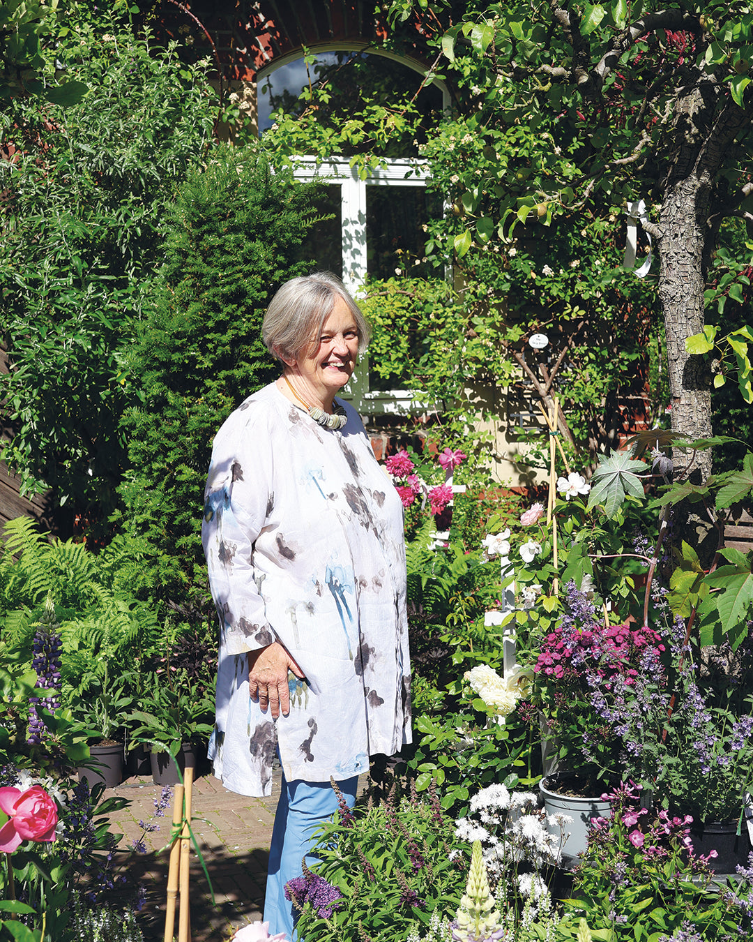 Eine lächelnde ältere Frau mit grauem Haar steht in einem lebendigen Garten, der die MASTER CLASS "Gestalten Sie Ihr eigenes Beet" von Dr. Isabelle Van Groeningen symbolisiert, die vom 19. bis 20. März 2026 stattfindet, mit Blumen und Grünzeug um sie herum und einem Backsteingebäude dahinter.