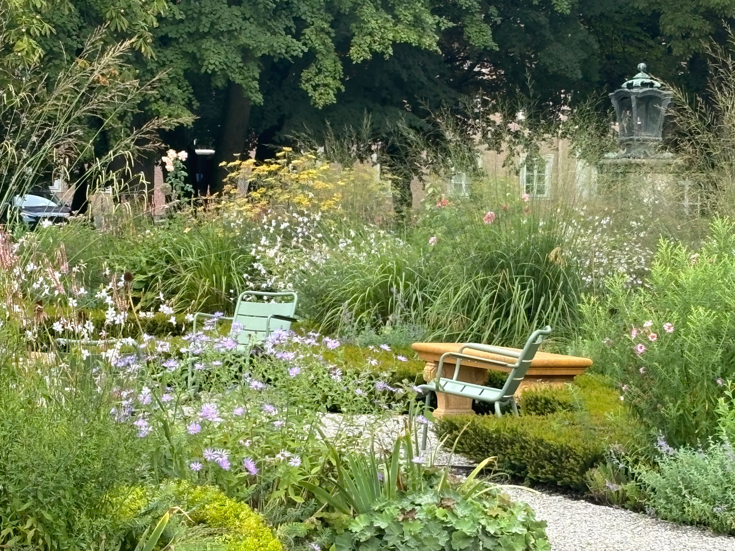 Zwei grüne Metallstühle und eine Bank stehen in einem üppigen Garten mit blühenden Pflanzen, Staudenbepflanzung und einem Kiesweg - ganz im Stil des Meisterkurses Gartenpflege der Königlichen Gartenakademie vom 03. bis 04.02.2026.