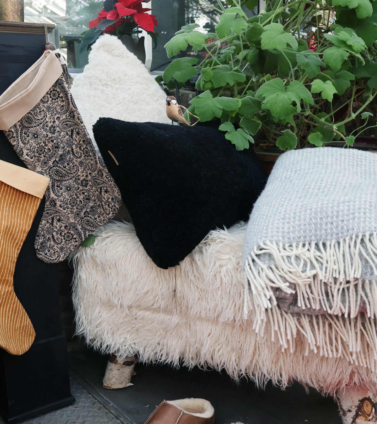 Eine gemütliche Bank mit dem Alice Kissenbezug aus Schafsfell in Creme (50x50) von Shepherd of Sweden, schwarze und weiße Kissen, eine graue Fransendecke, festliche Strümpfe, Grünpflanzen und Deko für echte skandinavische Gemütlichkeit.