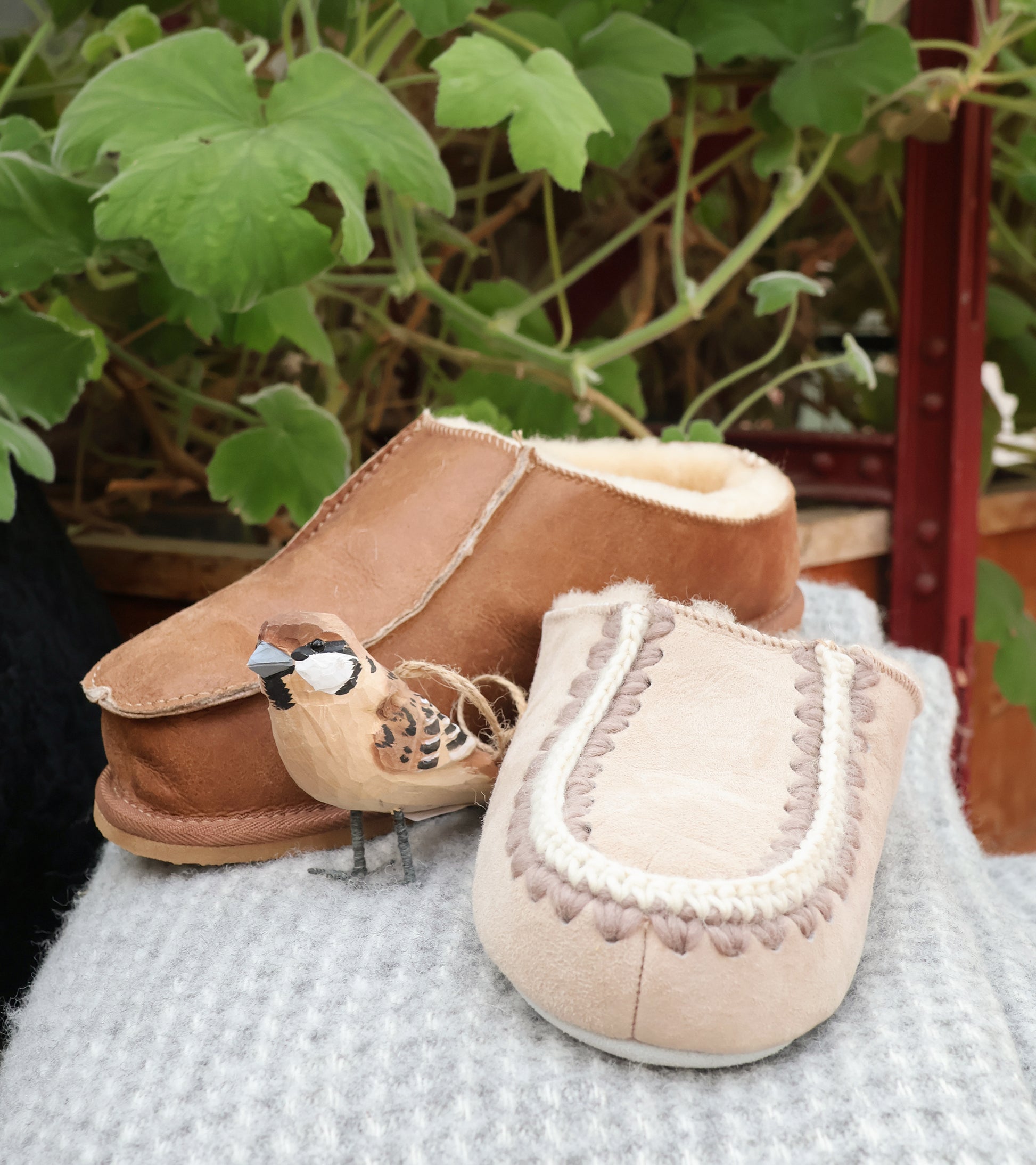 Zwei Shepherd of Sweden Beatrice Slipper aus Schaffell | Honey 39 Wollpantoffeln ruhen auf grauem Stoff. Vorne sitzt eine geschnitzte Vogelfigur, im Hintergrund stehen grüne Blattpflanzen, die eine gemütliche Atmosphäre schaffen.