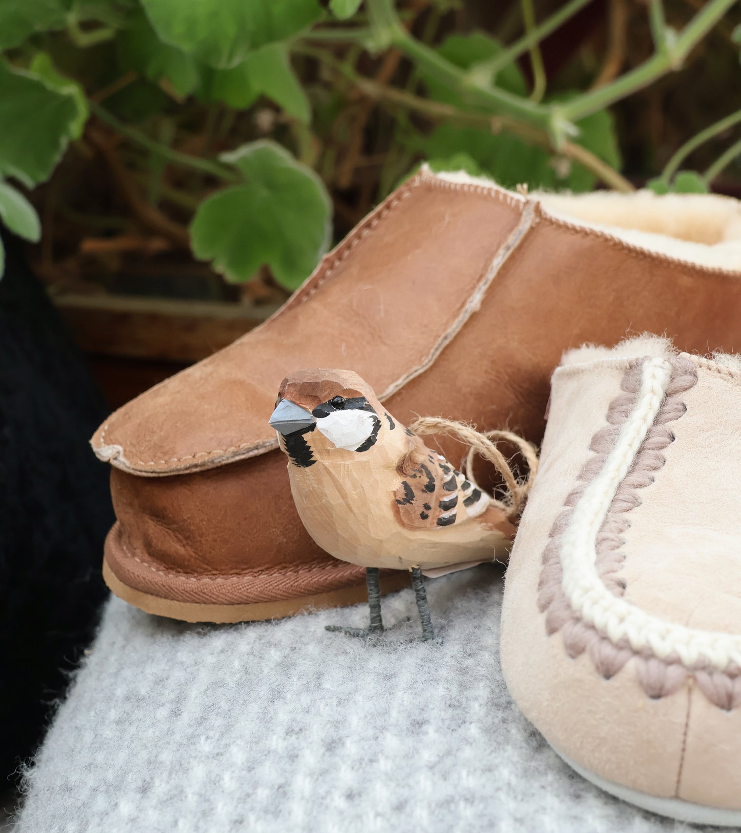 Eine kleine, detailreiche Vogelfigur aus Holz sitzt auf einer grauen Stofffläche zwischen zwei Shepherd of Sweden Richard Slipper aus Schaffell in Cognac (Größe 44), mit verschwommenen grünen Pflanzenblättern im Hintergrund.