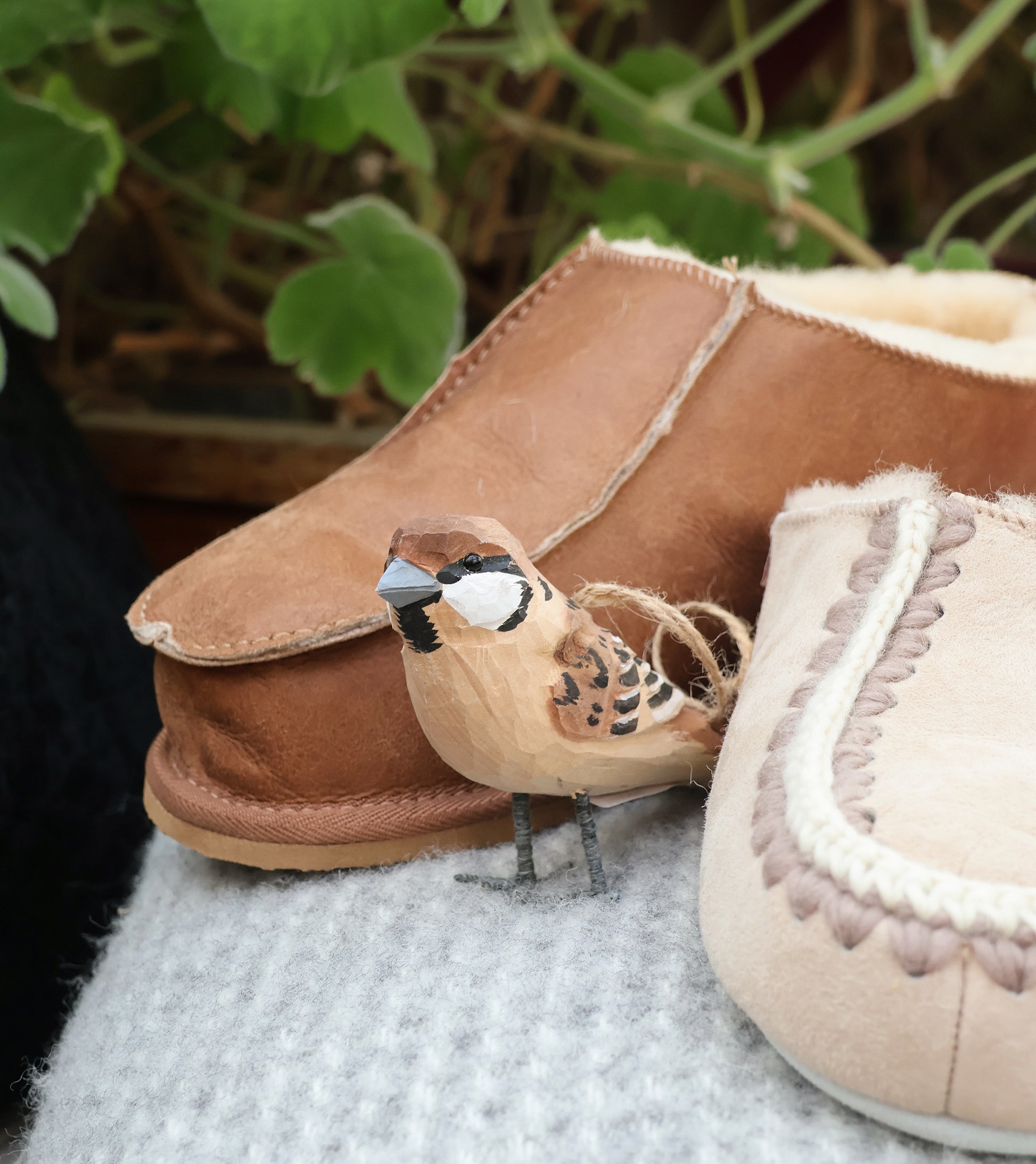 Eine kleine, detailreiche Vogelfigur aus Holz sitzt auf einer grauen Stofffläche zwischen zwei Shepherd of Sweden Richard Slipper aus Schaffell in Cognac (Größe 44), mit verschwommenen grünen Pflanzenblättern im Hintergrund.