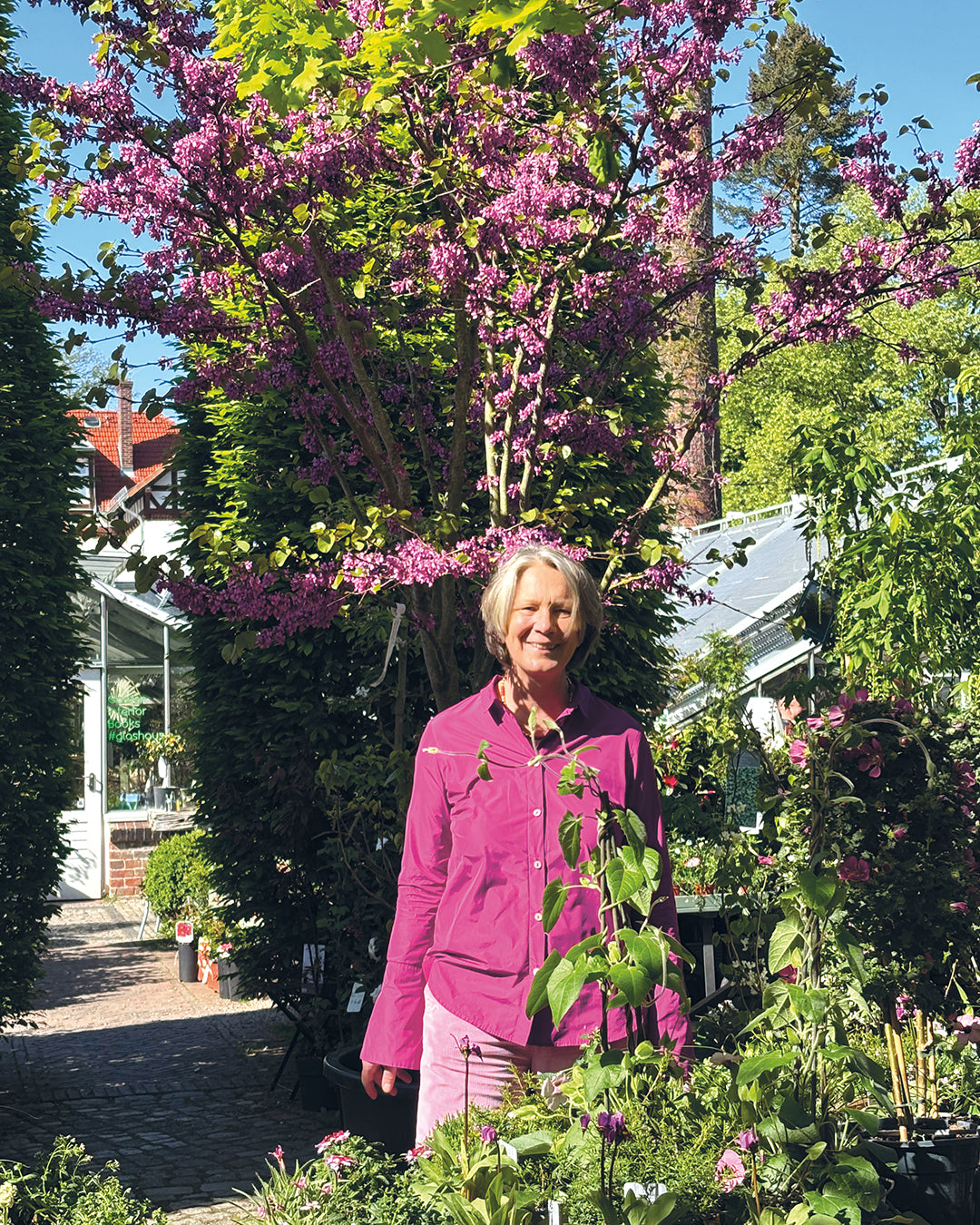 Eine Frau in einer rosafarbenen Jacke lächelt inmitten von blühenden Blumen und Grünzeug und genießt die Schönheit der Gartengestaltung beim "Do. 12.03.2026 | 10.00-13.00 HALBTAGESKURS Grundlage der Gartengestaltung" von Gabriella Pape.