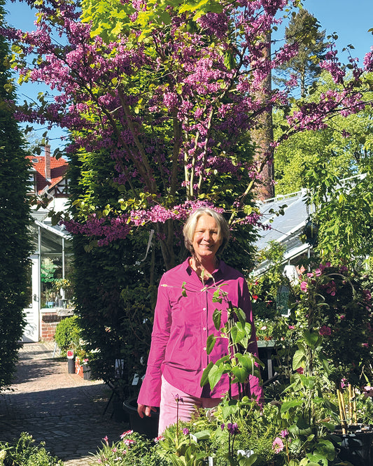 Eine Frau in einer rosafarbenen Jacke lächelt inmitten von blühenden Blumen und Grünzeug und genießt die Schönheit der Gartengestaltung beim "Do. 12.03.2026 | 10.00-13.00 HALBTAGESKURS Grundlage der Gartengestaltung" von Gabriella Pape.