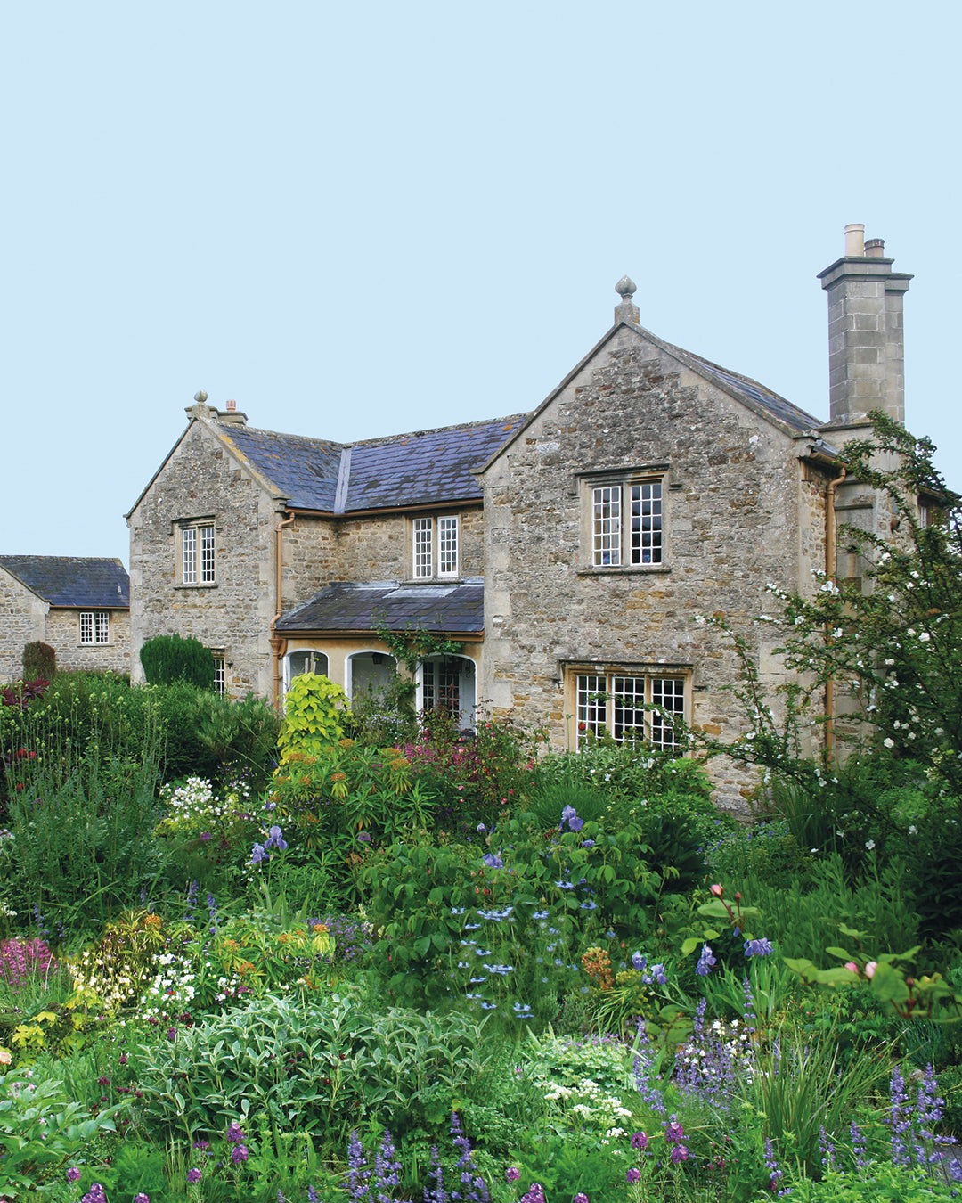 Erleben Sie den Charme eines Steinhäuschens inmitten von Rosen und üppigem Grün bei "Sa. 07.03.2026 | 13.30-16.30 HALBTAGESKURS Der Cottage Garten: Der Garten der Zukunft" von Dr. Isabelle Van Groeningen, umgeben von leuchtenden Blumen unter einem klaren Himmel.