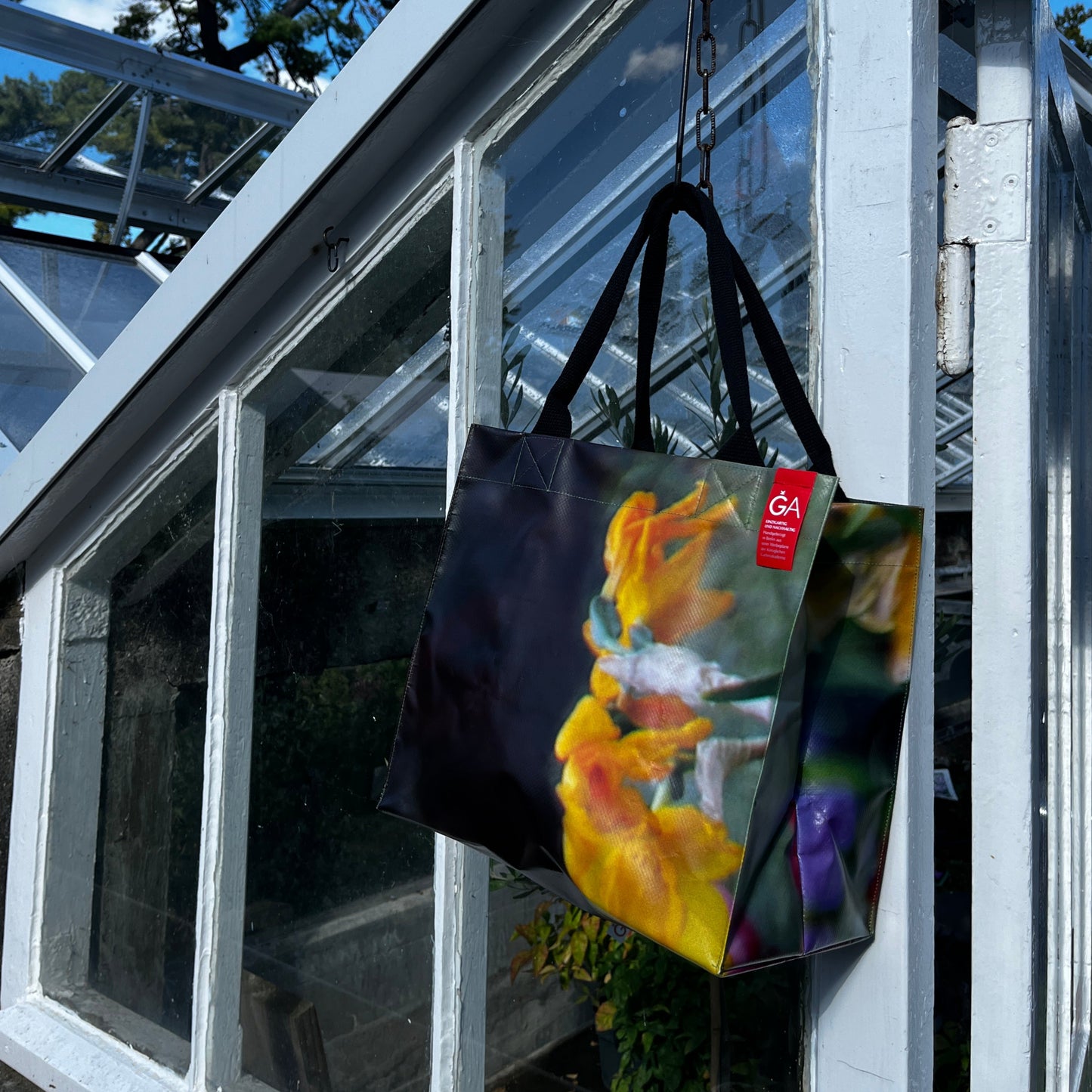 Die KGA Upcycling Tasche 'Narzisse' der Königlichen Gartenakademie mit buntem Blumenmuster hängt an einer gläsernen Gewächshaustür mit blauem Himmel und Baumzweigen im Hintergrund.