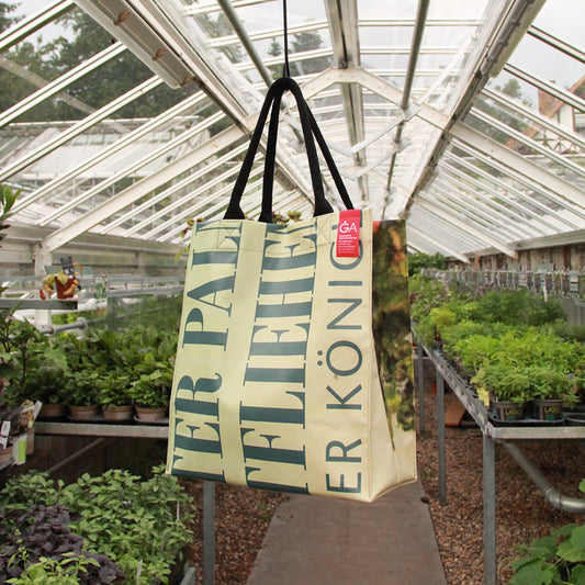 Der Upcycling KGA Shopper 'Orange' von der Königlichen Gartenakademie mit frechem grünem Schriftzug hängt von der Decke in einem mit Pflanzen gefüllten Gewächshaus, während das Sonnenlicht durch das Glasdach strömt.