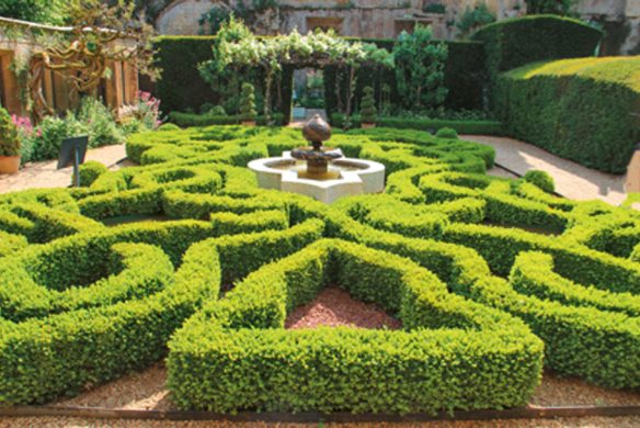 Gestalten Sie, inspiriert von "Gärtnern wie Gott in England" aus dem Callwey Verlag, einen formalen Garten mit symmetrischen Buchsbaumhecken, einem zentralen Steinbrunnen, Kieswegen und üppigem Grün im Stil der englischen Gartenkunst.