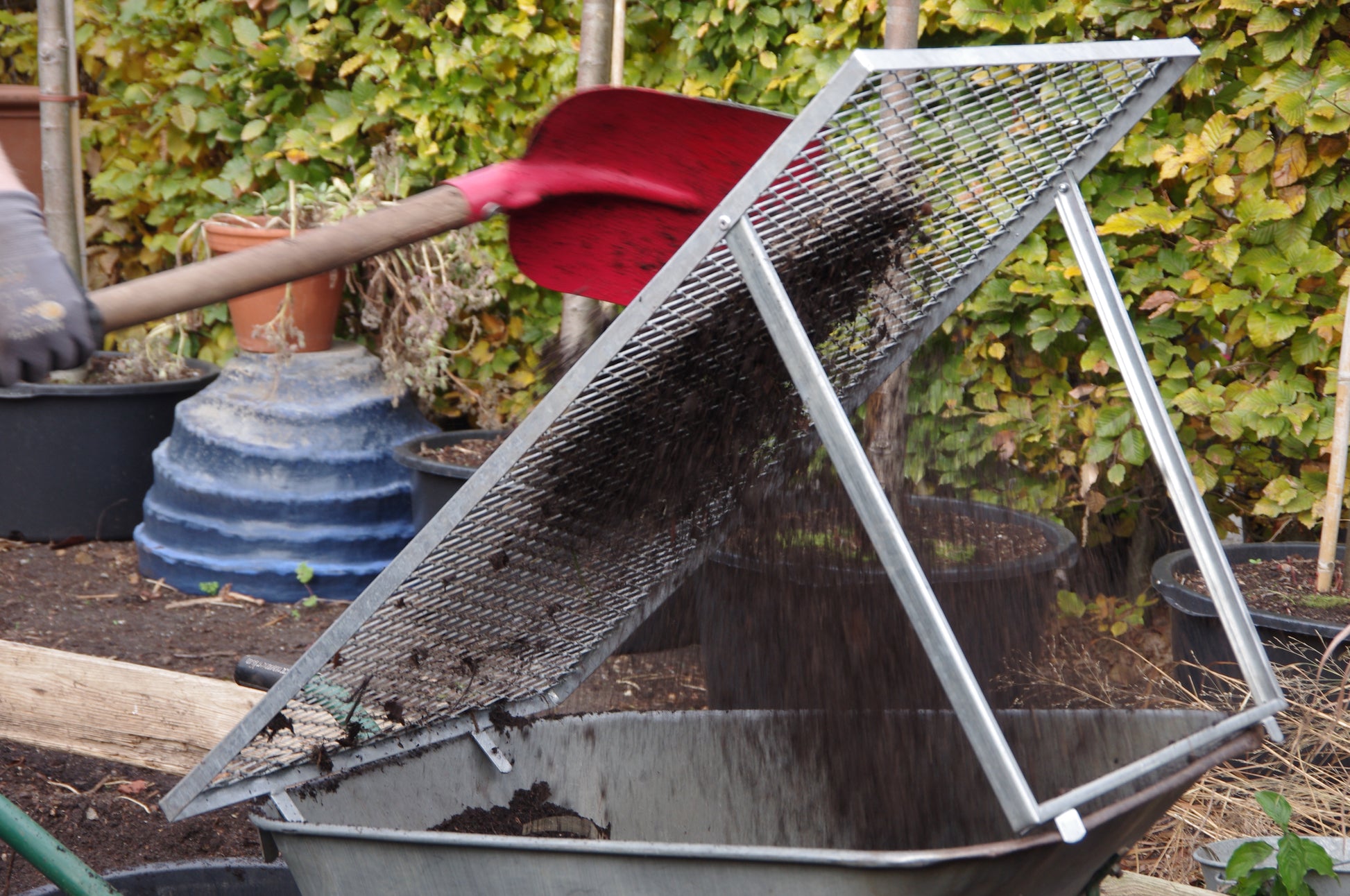 In Thea Carlins "WORKSHOP Kompost: Das wahre Gold des Gärtners" siebt eine Person mit einer roten Schaufel Erde und Kompost durch ein Netz in eine Schubkarre; im Hintergrund erscheinen Gartentöpfe, Grünzeug und Bodenaktivator.