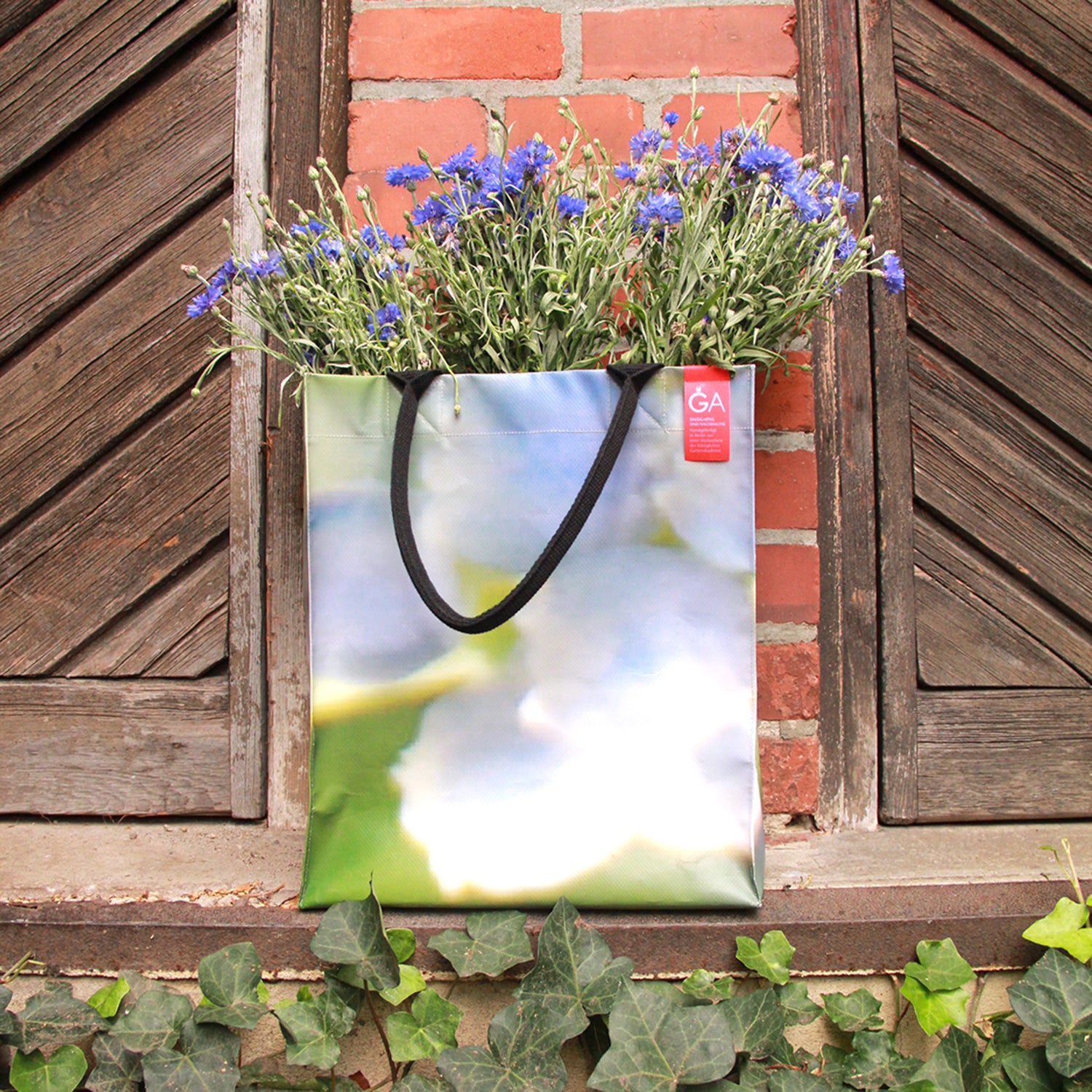 Die Upcycling-Tasche 'Leaf' aus Werbeplane von Königliche Gartenakademie mit verschwommenem Blumenmuster hängt an einem rustikalen Fenster und ist mit lila Wildblumen gefüllt, die vor dem Hintergrund von Ziegelsteinen und Efeu Gartencharme versprühen.