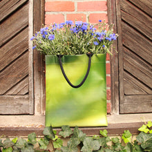 Lade das Bild in den Galerie-Viewer, Ein grüner Upcycling KGA Shopper &#39;Leaf&#39; von der Königlichen Gartenakademie, gefüllt mit violetten Blumen, steht auf einem Fenstersims mit verwitterten Fensterläden, einer Backsteinmauer und grünem Efeu darunter.
