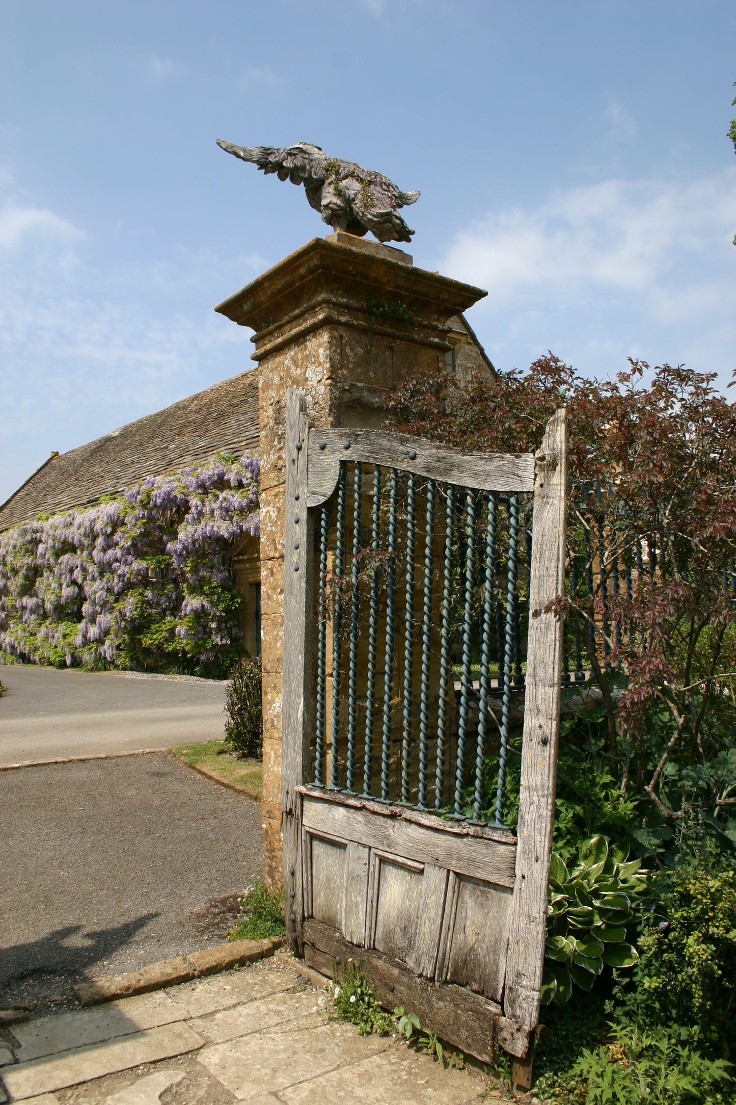 Ein verwittertes Holztor an einer Steinsäule mit einer Vogelskulptur, umrahmt von lila Blumen, erinnert an den Charme von Südengland - perfekt für Dr. Isabelle Van Groeningens Afternoon-Tea-Vortrag: Die Gärten von Somerset, Dorset und Wiltshire.