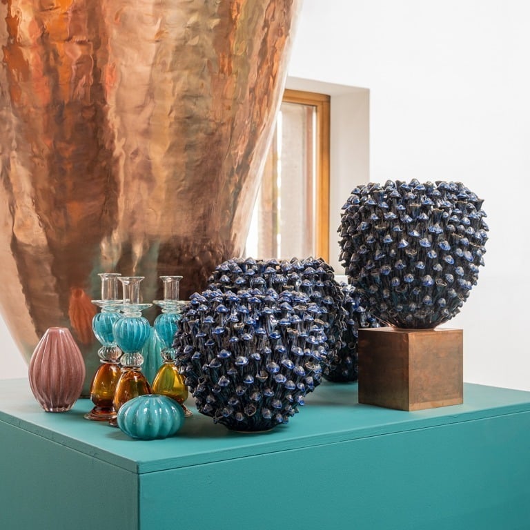 Ein tealfarbener Sockel präsentiert die Des Pots Vase | Blau skulptural L, gepaart mit gerippten Glasflaschen in Aqua und Rosa, bernsteinfarbenen Behältern sowie einer großen Kupferskulptur und einem Fenster im Hintergrund.