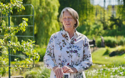 Eine Frau steht in einem sonnenbeschienenen Garten, hält ihr Smartphone in der Hand und trägt eine geblümte Bluse. Sie scheint sich für den "HALBTAGESKURS Grundlagen der Gartengestaltung" von Gabriella Pape an der Königlichen Gartenakademie am 06.11.2025 zu interessieren.