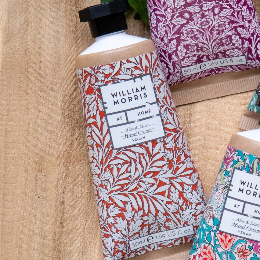 Eine Nahaufnahme der William Morris at Home | 'Foliage' Handcreme 50 ml Tuben mit komplizierten floralen Mustern, die auf Holz liegen. Diese vegane Formel enthält den pflegenden Duft von Aloe und Limette von William Morris at Home.