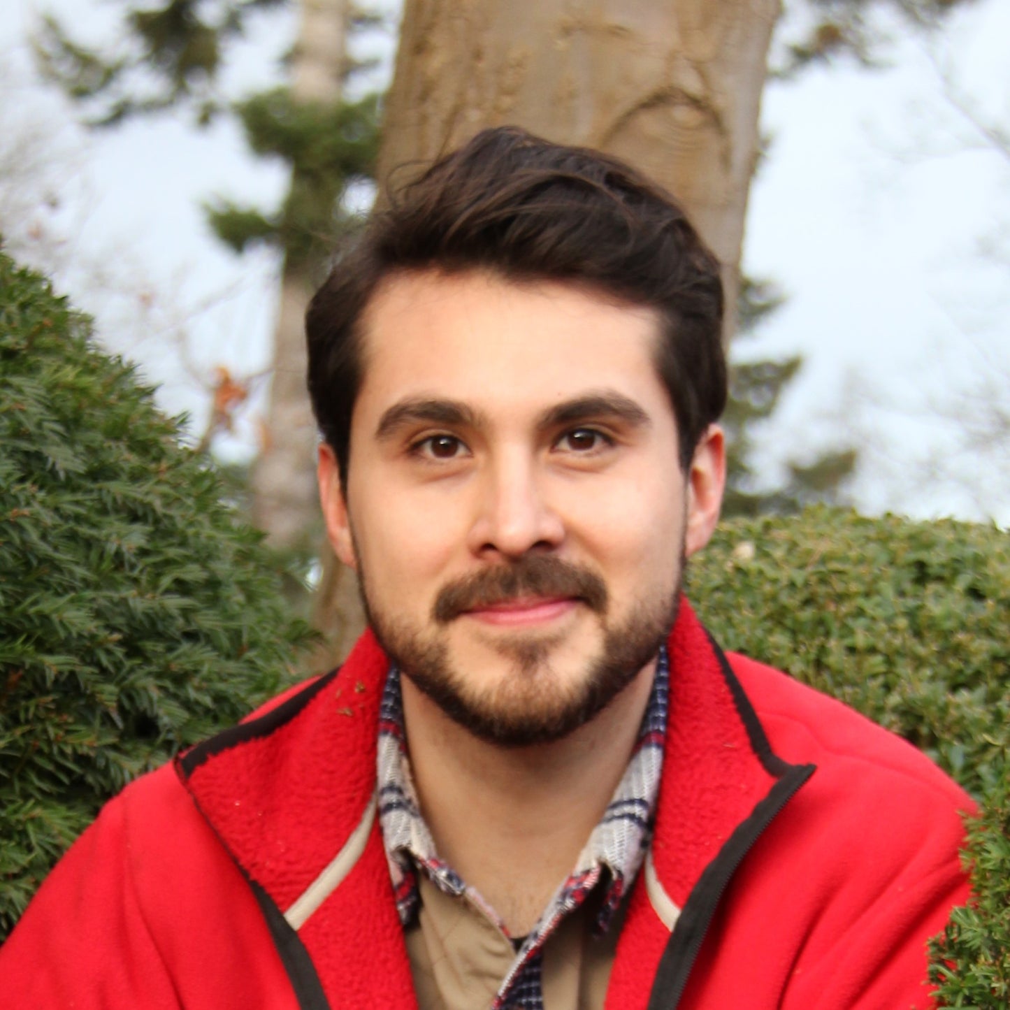 Ein junger Mann mit dunklem Haar und Bart, in roter Jacke und kariertem Hemd, lächelt im Freien zwischen grünen Sträuchern und erfreut sich an dem üppigen Wachstum, das von Sebastian Keller-Lewis' WORKSHOP Glücklicher Boden, glückliche Pflanzen am 27.02.2026 inspiriert wurde.