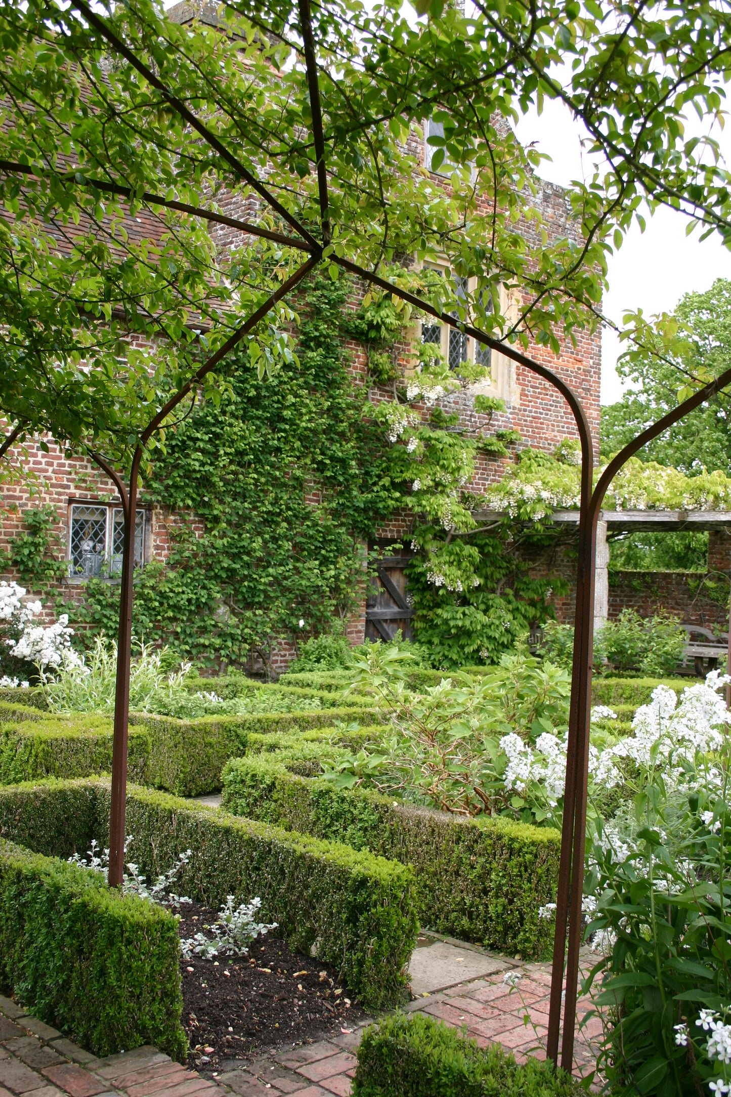 Inspiriert von Dr. Isabelle Van Groeningens "Die Gartenpracht von Kent und Sussex", zeigt dieser üppige Garten Buchsbaumhecken in geometrischen Mustern, leuchtende Blüten, eine Metalllaube und ein mit Efeu bedecktes Backsteingebäude mit weißen Blumen.