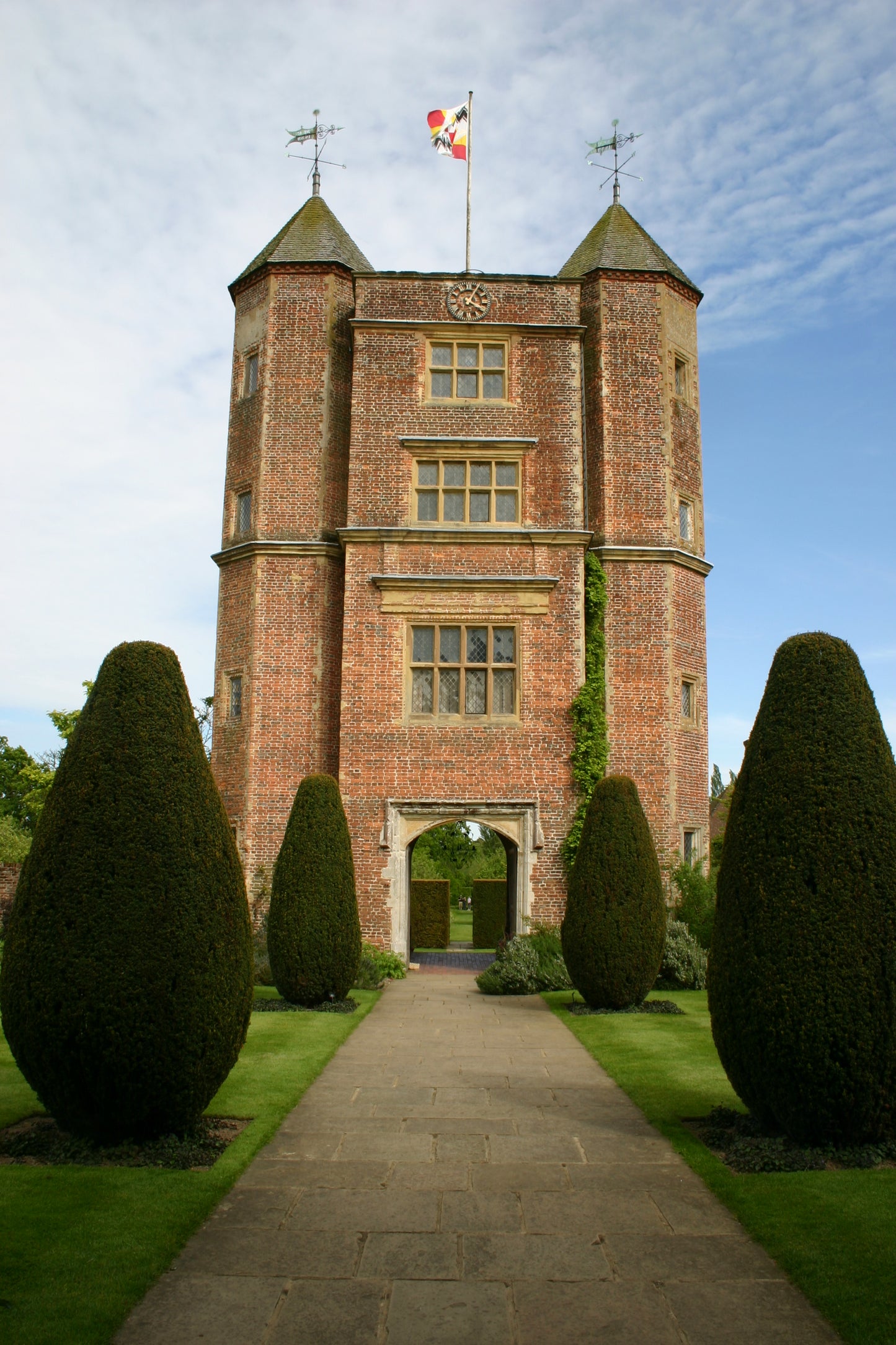 Ein hoher Backsteinturm, flankiert von gepflegten Sträuchern und Rasenflächen wie Gärten Kent Sussex, steht unter einem teilweise bewölkten Himmel - erleben Sie diese Eleganz beim AFTERNOON-TEA-VORTRAG von Dr. Isabelle Van Groeningen am 17.02.2026, 14.30-15.30 Uhr.