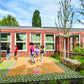 Kinder spielen gerne draußen auf einer gepflasterten Fläche neben einem modernen Backsteingebäude mit großen Fenstern und Grünflächen, das an einem sonnigen Tag das Ergebnis der durchdachten Gestaltung von Landschaftsarchitektur 2025 des Callwey Verlags zeigt.