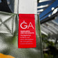 Eine Nahaufnahme eines roten Etiketts auf der grünen Upcycling Tasche 'Gewächshaus' aus Werbeplane von Königliche Gartenakademie. Auf dem Etikett steht: GA. Einzigartig und nachhaltig. Handgefertigt in Berlin aus einer Werbeplane der Königlichen Gartenakademie.