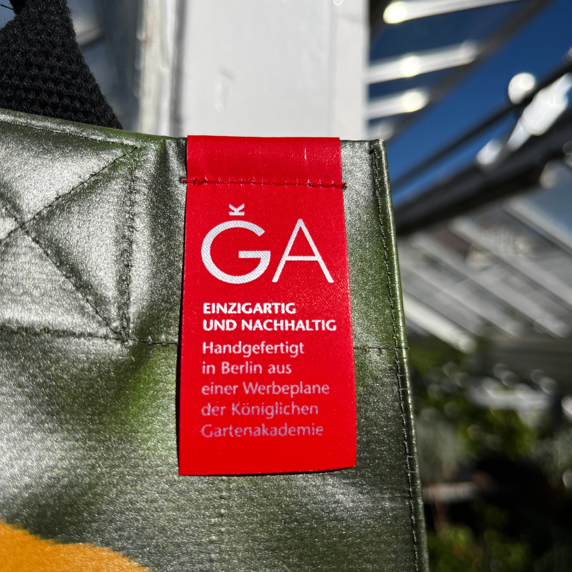 Eine Nahaufnahme der KGA Upcycling Tasche "Gewächshaus" der Königlichen Gartenakademie zeigt ein rotes Schild mit der Aufschrift "KGA Einzigartig und nachhaltig. Handgefertigt in Berlin aus einer Werbeplane." Das Sonnenlicht hebt das Schild hervor.