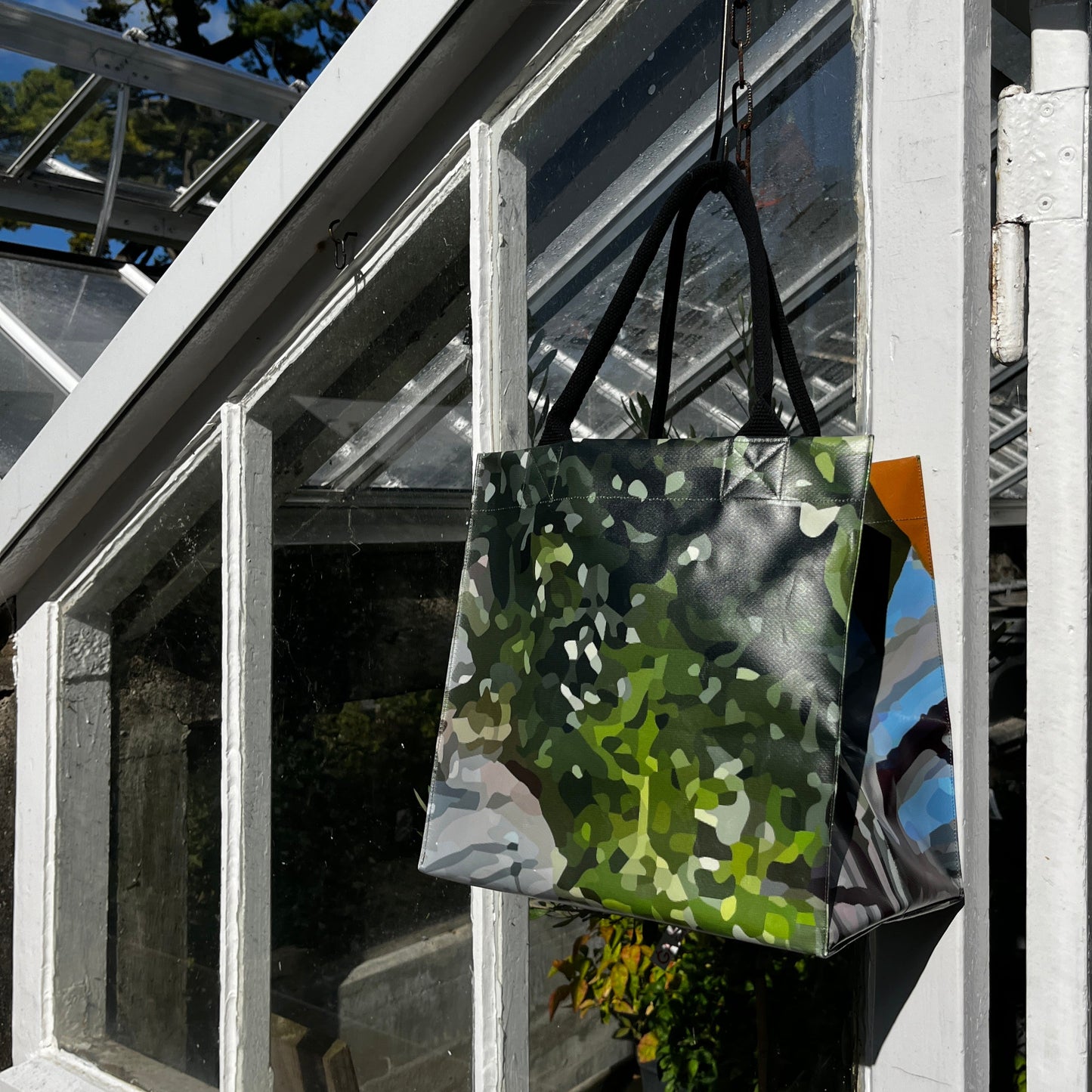 Die grün-schwarz gemusterte KGA Upcycling Tasche "Gewächshaus" von der Königlichen Gartenakademie hängt an einer weißen Gewächshaustür, durch deren Glas das Sonnenlicht fällt und hinter der das Grün des Gartens zu sehen ist.
