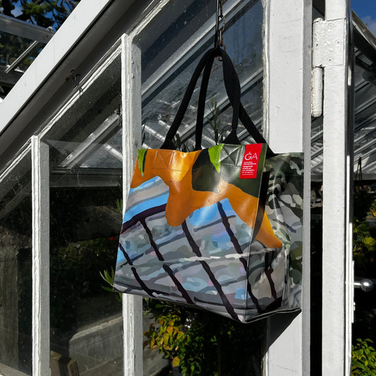Die KGA Upcycling Tasche "Gewächshaus" der Königlichen Gartenakademie mit floralem Design hängt an einem Gewächshaustürgriff, so dass Sonnenlicht und Pflanzen im Inneren sichtbar sind.