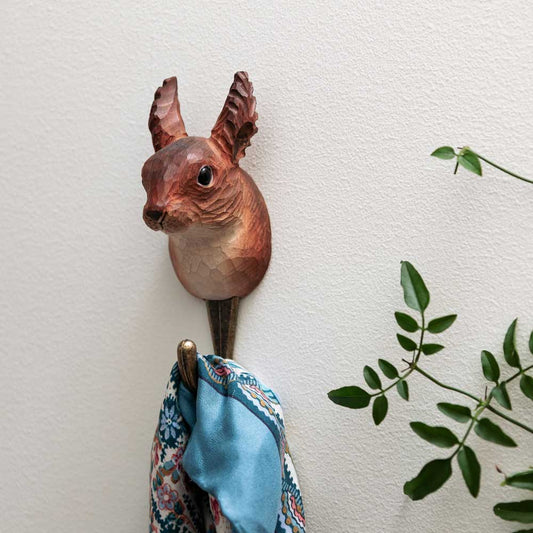 Der handgeschnitzte Wandhaken aus Holz "Eichhörnchen" von Wildlife Garden hält einen blau gemusterten Schal, daneben eine grüne Pflanze vor weißer Wand - ein originelles Geschenk für Natur- und Tierfreunde.