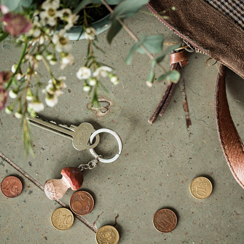 Ein Schlüsselset mit dem handgefertigten Schlüsselanhänger aus Holz | Steinpilz von Wildlife Garden liegt auf Stein, umgeben von Münzen, einer Pflanze und einer braunen Ledertasche - ein ideales Geschenk für Naturfreunde.