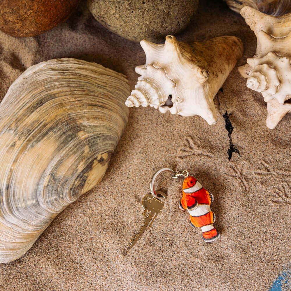 Ein handgefertigter Schlüsselanhänger aus Holz | Clownfisch von Wildlife Garden liegt auf sandigem Boden neben Muscheln und Steinen - ideal als Geschenk für Meereswelt-Fans.