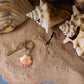 Ein handgefertigter Schlüsselanhänger aus Holz | Muschel von Wildlife Garden liegt auf sandigem Boden neben Muscheln und Vogelspuren.