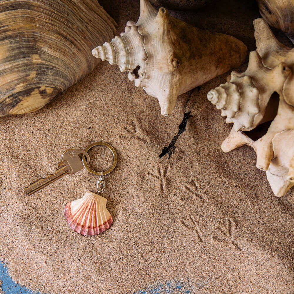Ein handgefertigter Schlüsselanhänger aus Holz | Muschel von Wildlife Garden liegt auf sandigem Boden neben Muscheln und Vogelspuren.