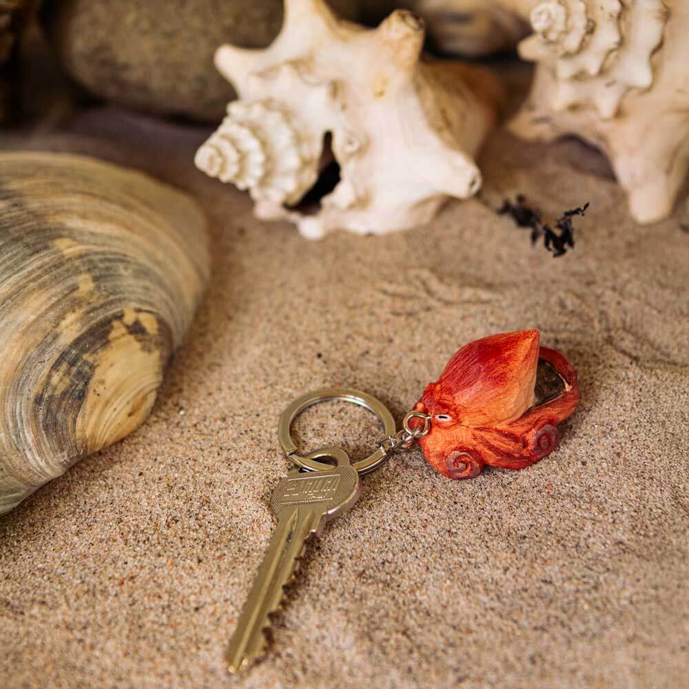 Ein handgefertigter Schlüsselanhänger aus Holz | Oktopus von Wildlife Garden liegt auf sandigem Grund zwischen Muscheln - ein ideales maritimes Geschenk.