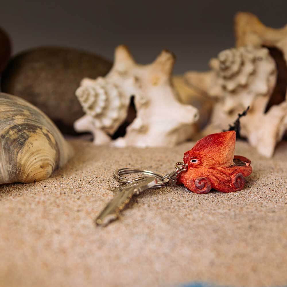 Der handgefertigte Schlüsselanhänger aus Holz "Oktopus" von Wildlife Garden hält einen silbernen Schlüssel und liegt auf sandigem Boden, umgeben von Muscheln und Steinen.