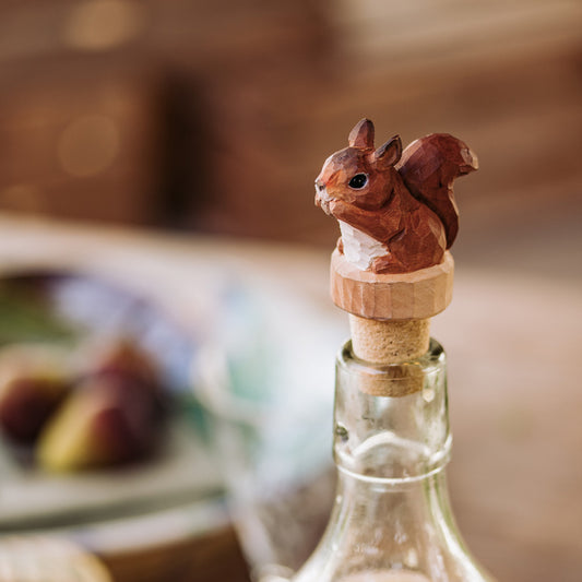 Der handgeschnitzte Bottle Stopper Squirrel von Wildlife Garden aus Holz sitzt auf einer Flasche; der sanft verschwommene Hintergrund zeigt ein Tischgedeck - ideal als charmantes Geschenk.