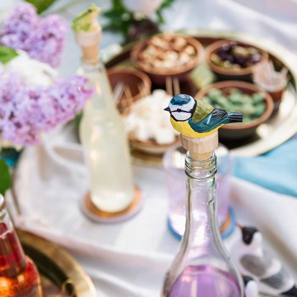 Ein handgeschnitzter Flaschenverschluss aus Holz in Blaumeise-Form von Wildlife Garden schmückt eine Glasflasche auf einem festlich gedeckten Tisch mit Drinks, Blumen und Snacks - ein farbenfrohes Geschenk für besondere Anlässe.