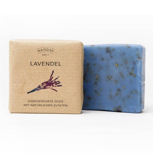Ein Riegel maisoap berlin - Lavendel Seife mit sichtbaren Lavendelstücken steht neben seiner braunen Papierverpackung mit der Aufschrift LAVENDEL, mit Lavendelkunst und deutschem Text - ideal für ein entspanntes Duscherlebnis und sanfte Hautpflege.