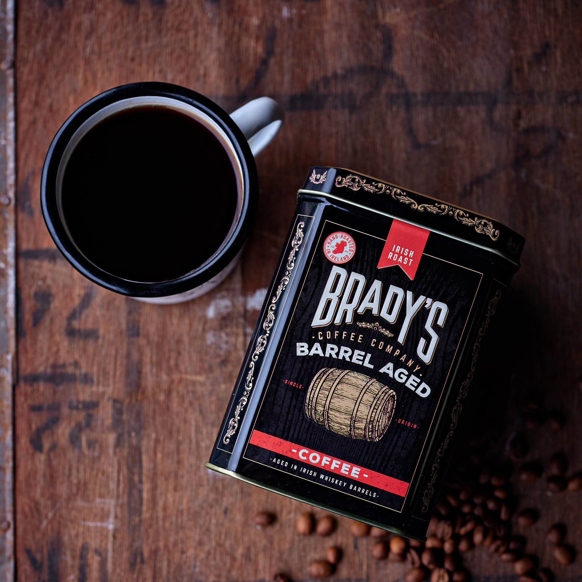 Eine Tasse schwarzen Kaffee steht neben Brady's Coffee Company – Brady's Kaffeedose mit ganzen Bohnen, im Fass gereift, irischer Whiskey. Ganze Bohnen dieses im Fass gereiften irischen Whiskeys sind auf der Holzoberfläche verstreut.