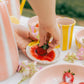 Eine Hand taucht eine frische Erdbeere in rote Soße auf dem Alfar Tierra Cocida | Kleiner Keramikreibe Teller ZIMT, umgeben von rosa und gelb gestreiften Tassen, Erdbeerschalen und rosa Blumen auf einem weißen Tisch.