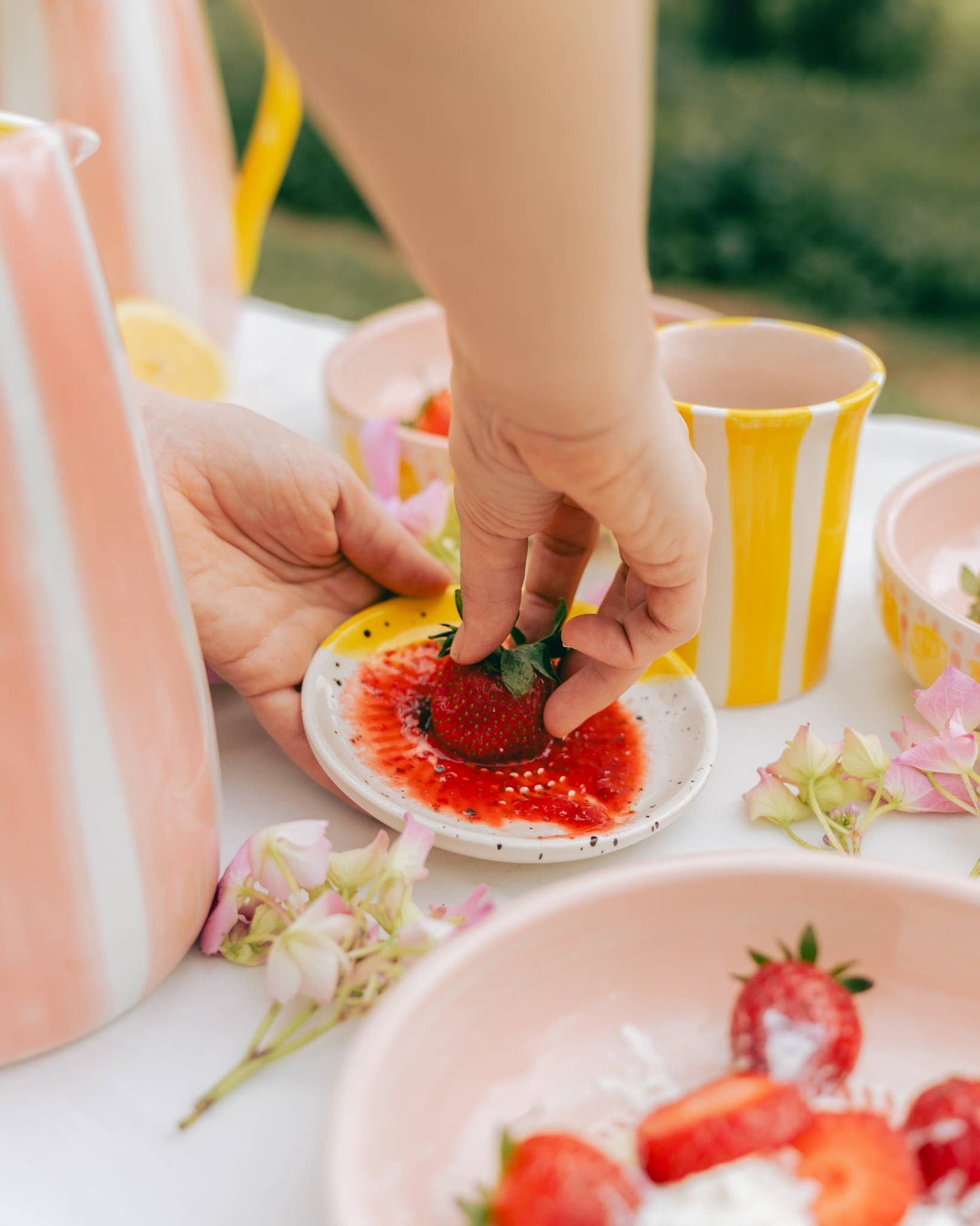 Eine Hand taucht eine frische Erdbeere in rote Soße auf dem Alfar Tierra Cocida | Kleiner Keramikreibe Teller ZIMT, umgeben von rosa und gelb gestreiften Tassen, Erdbeerschalen und rosa Blumen auf einem weißen Tisch.
