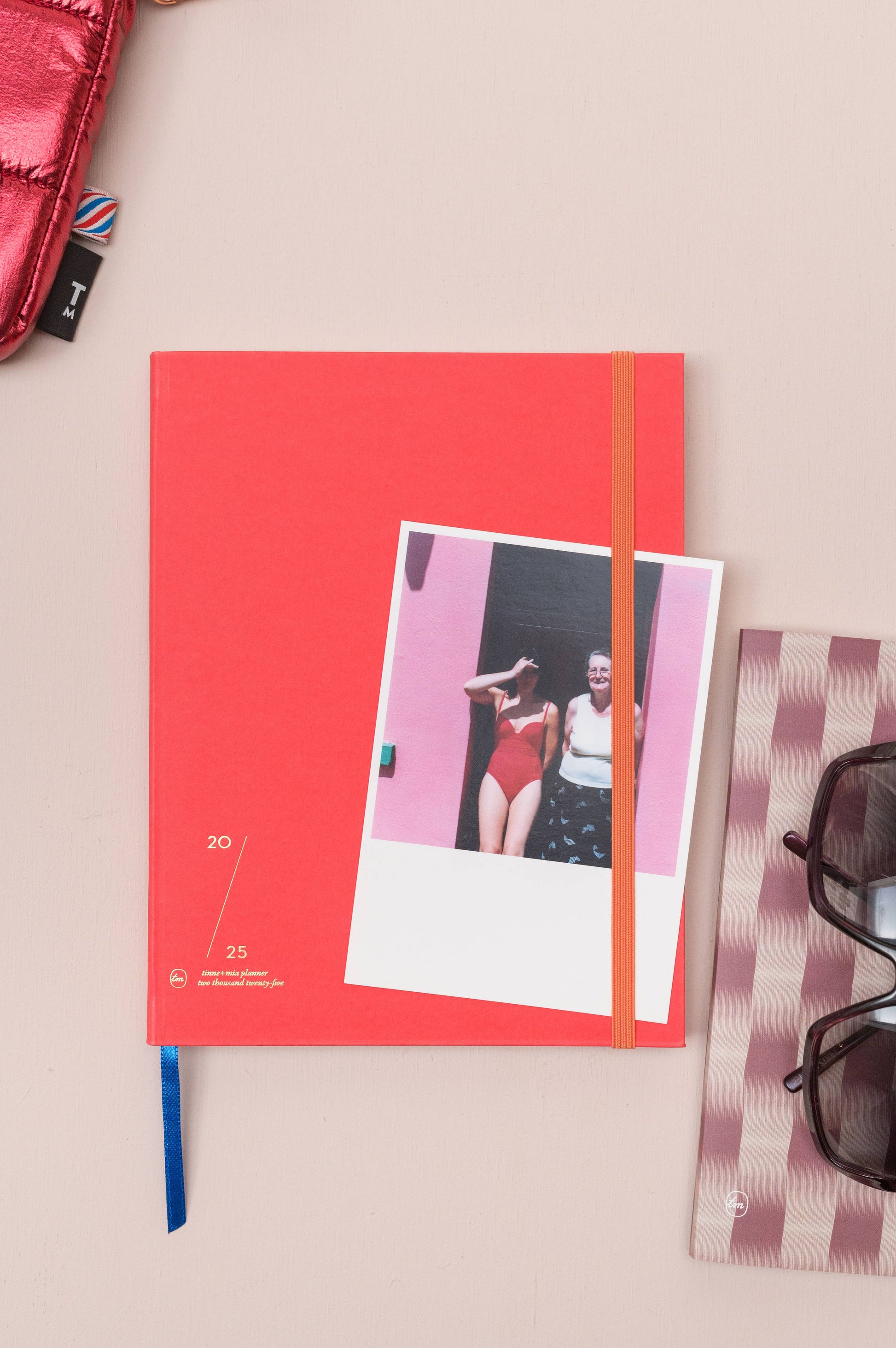 Ein Tinne Mia - Notizbuch mit japanischem Verschluss in Cinnamon von Tinne Mia liegt auf einer beigen Fläche, unter dessen Verschluss sich ein Polaroid eines lächelnden Paares befindet. In der Nähe befinden sich eine Sonnenbrille und ein rosa Metall-Accessoire.